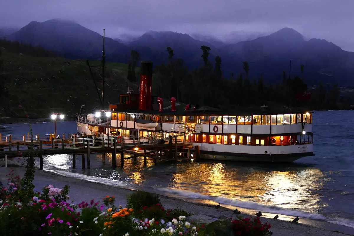 The TSS Earnslaw moored at the Walter Peak jetty at dusk, her windows lit, with a red funnel, hills behind and flowers in the foreground