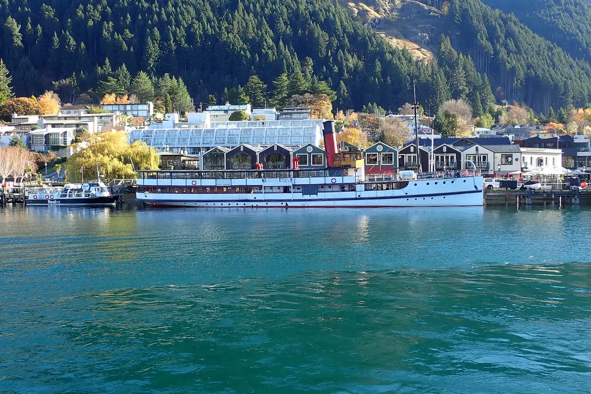 The white-hulled TSS Earnslaw with her red funnel moored at Steamer Wharf in Queenstown, with wooded hills and waterfront buildings behind