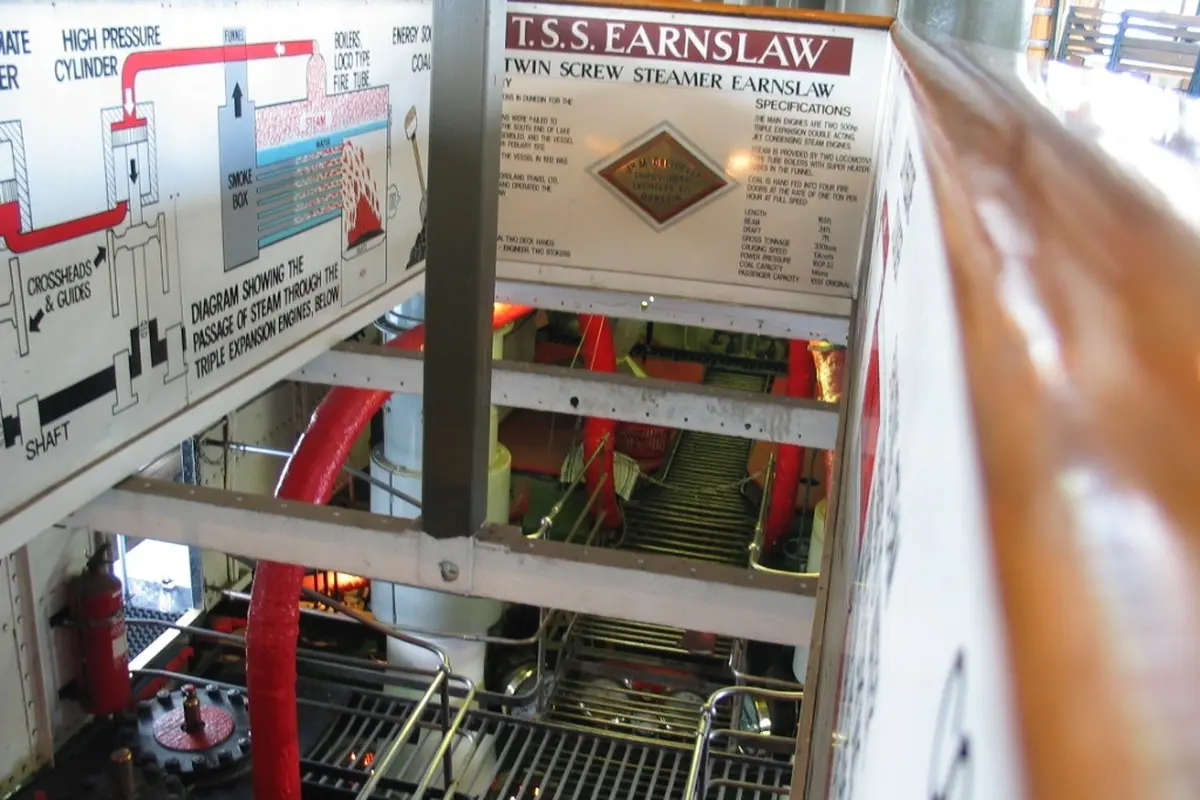 Looking down from a walkway into the TSS Earnslaw's engine room, past a signboard with a diagram of steam passing through triple-expansion engines