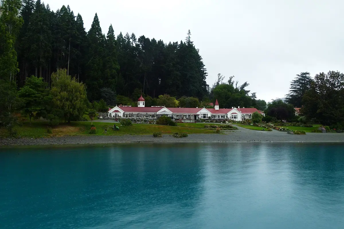 The red-roofed white homestead at Walter Peak High Country Farm seen across blue lake water, with dark conifers behind and gardens in front